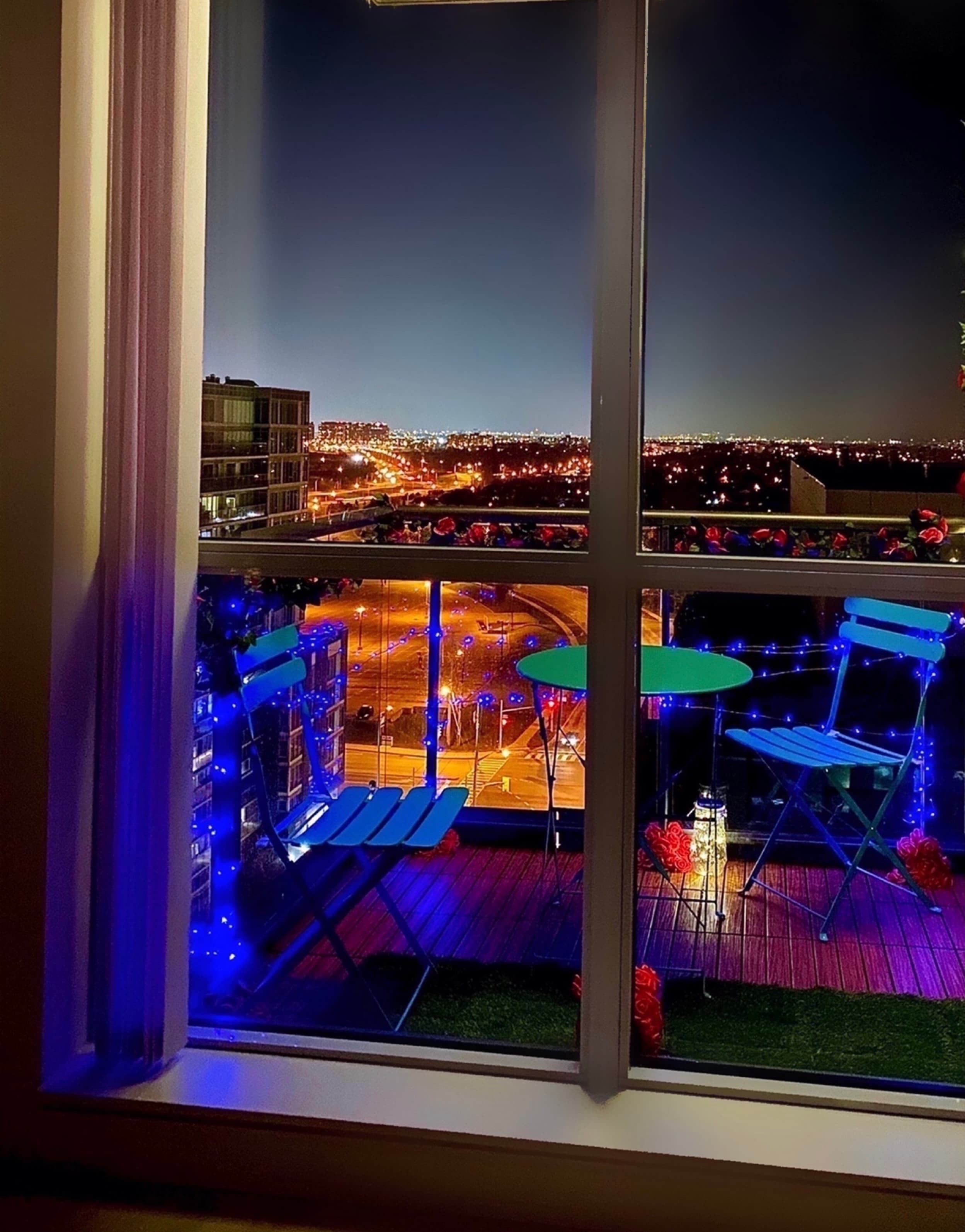 Balcony decorated with blue lights and bistro set overlooking a city skyline at night.