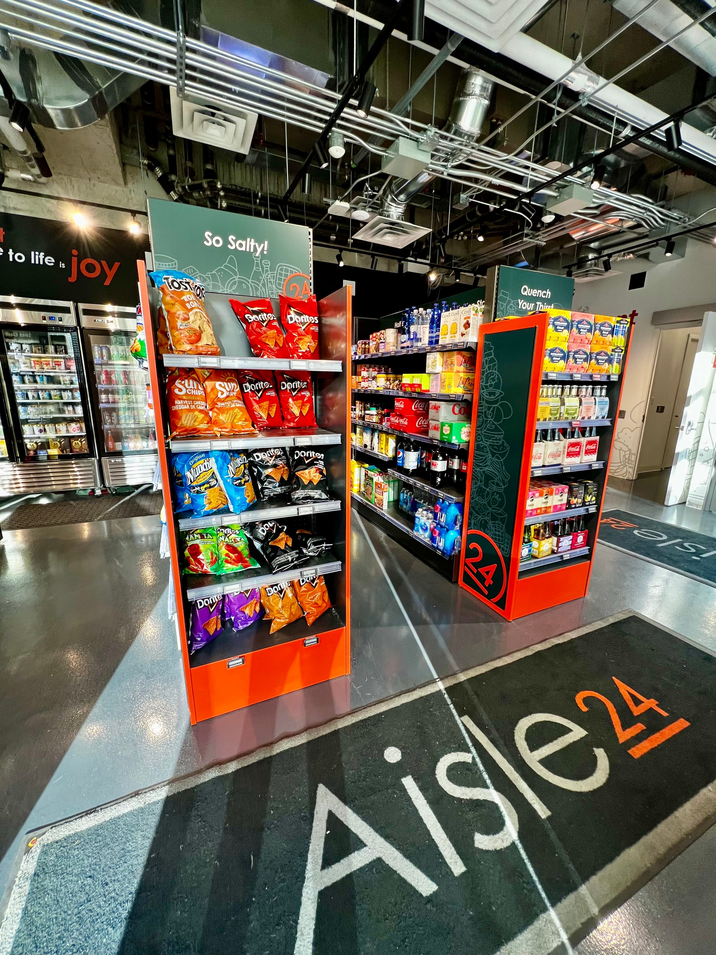 Shelves of snacks and drinks inside a modern Aisle 24 convenience store with industrial ceilings.