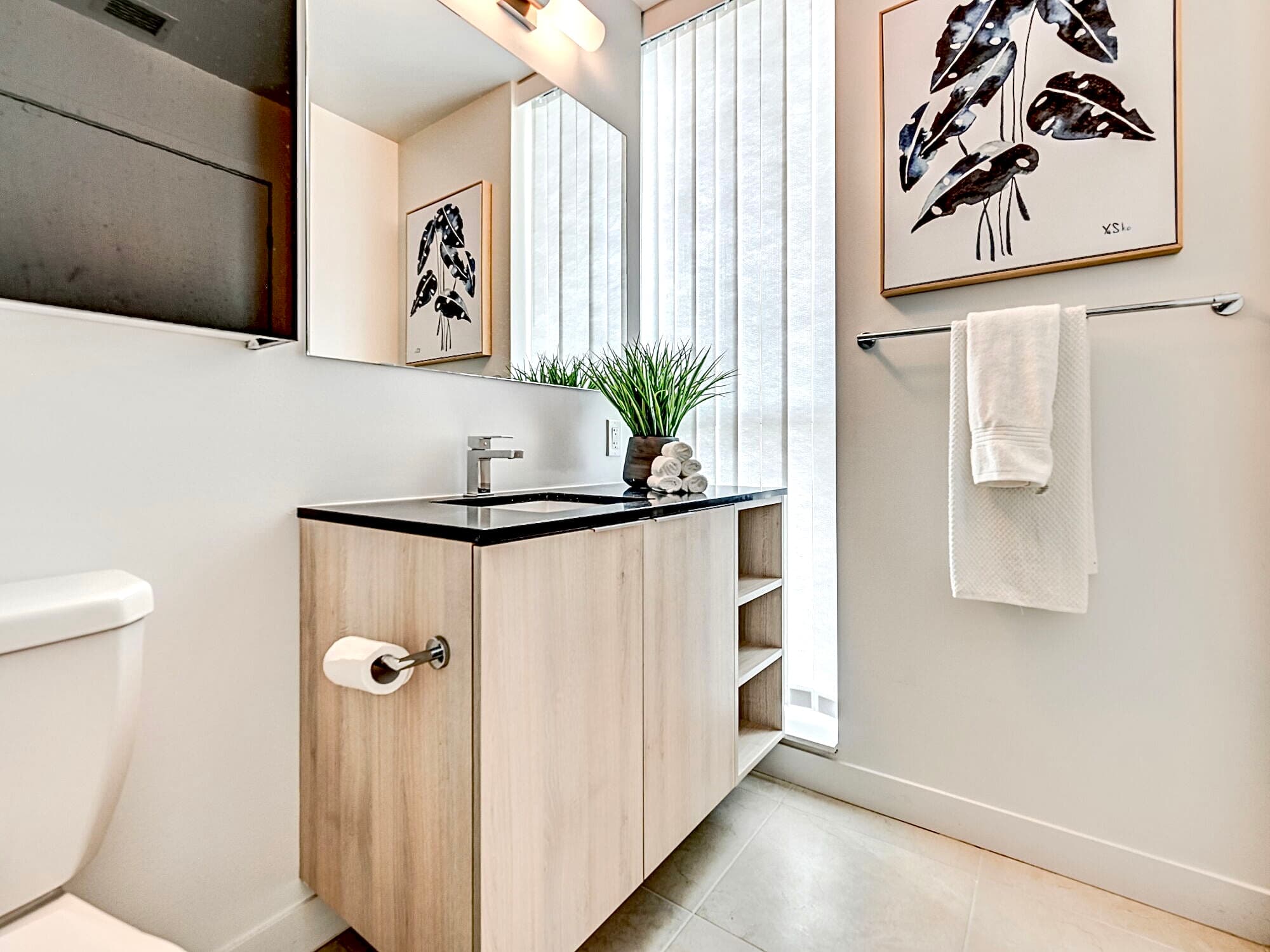 Modern bathroom with light wood vanity, black countertop, large mirror, and framed leaf art.