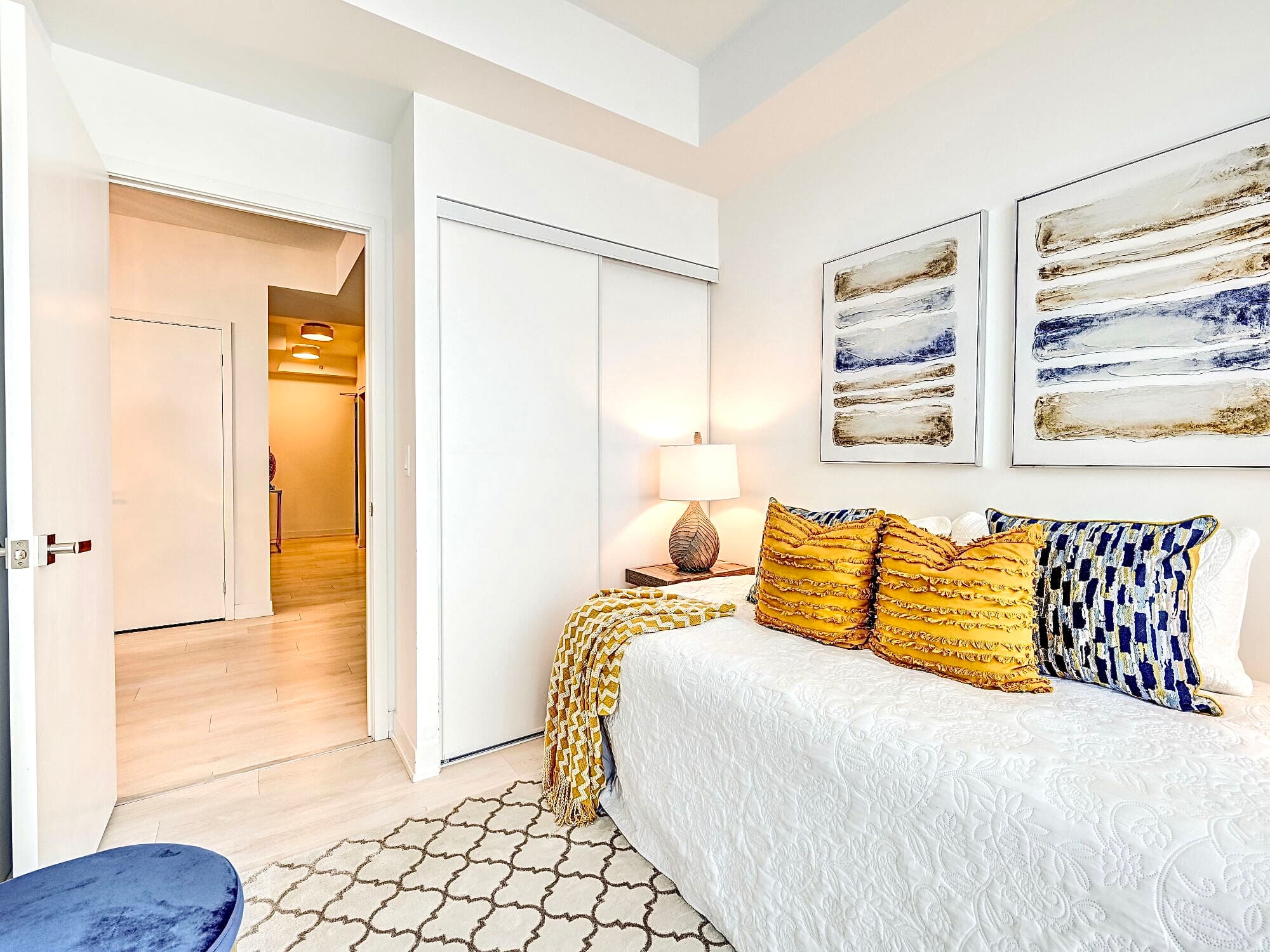 Modern bedroom with white bedding, yellow textured pillows, abstract art, and sliding closet doors.