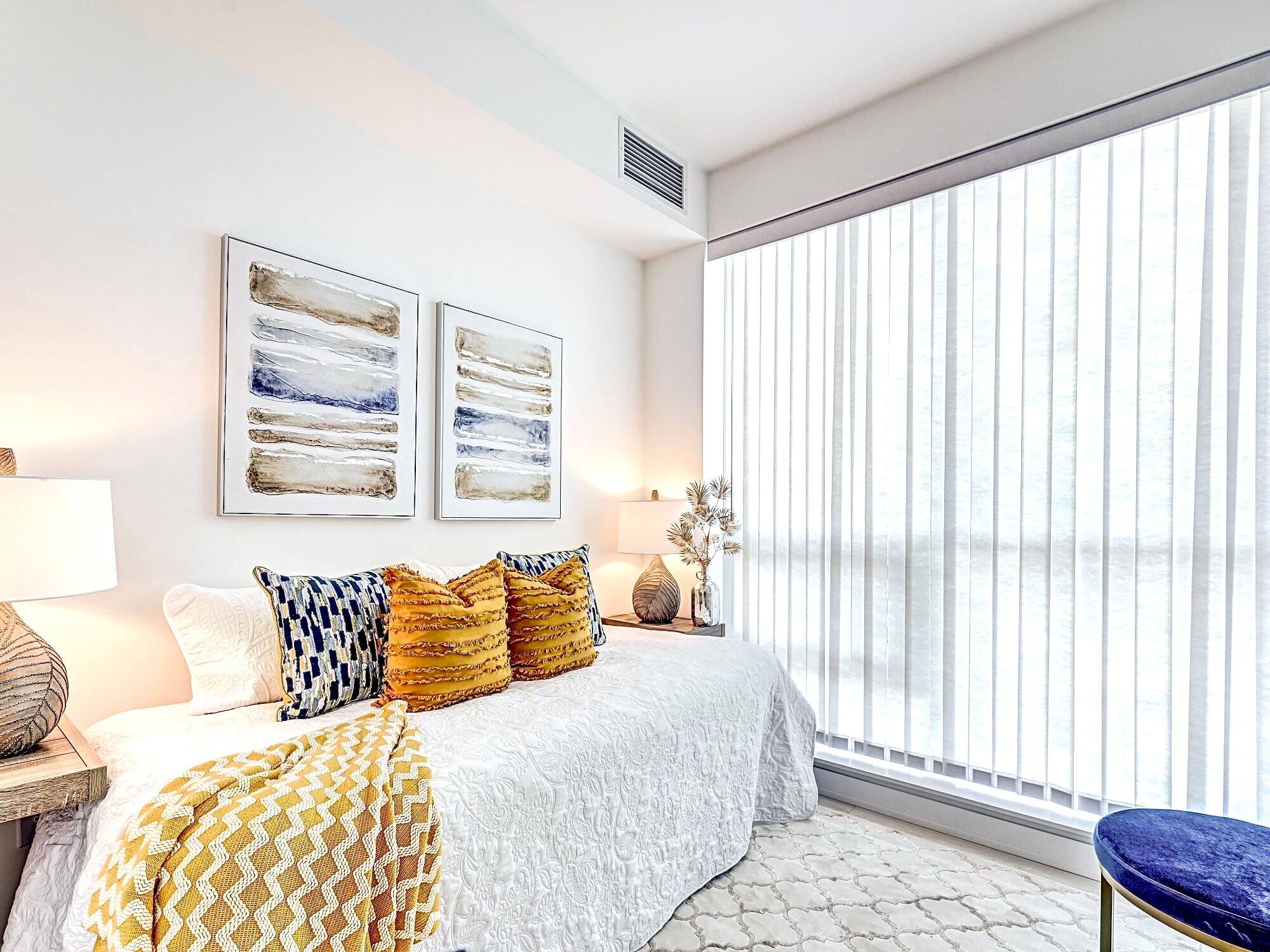 Bright bedroom with white bedding, yellow and blue pillows, abstract art, and large vertical blinds.