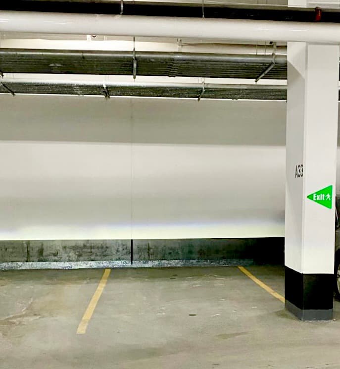 Empty parking space in a garage with white walls, overhead pipes, and green exit sign.